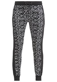 Zwarte broek met een geometrisch wit patroon, een elastische tailleband met trekkoord en effen zwarte zijpanelen. Ribgebreide boorden maken het ontwerp af.