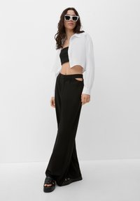 Zwarte cropped bandeau top, oversized witte button-up, zwarte wijde broek met uitsnijdetails, gedragen met chunky zwarte sandalen en witte zonnebril.