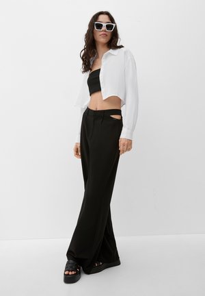 Czarny krótki top bandeau, oversize'owa biała koszula na guziki, czarne szerokie spodnie z wycięciami, zestawione z masywnymi czarnymi sandałami i białymi okularami przeciwsłonecznymi.
