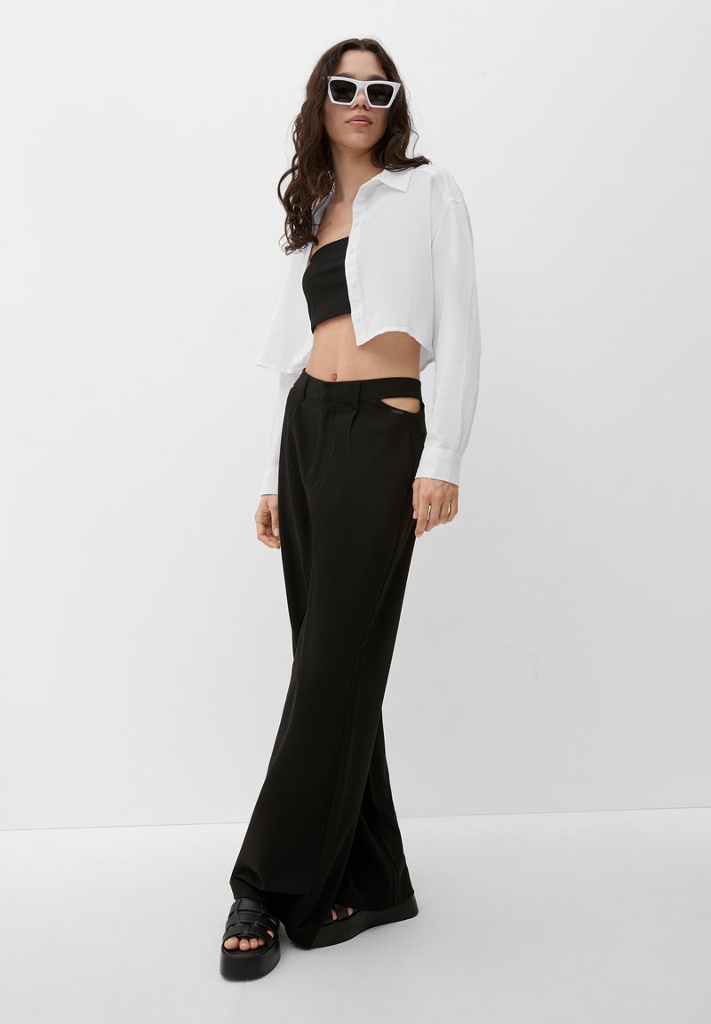 Zwarte cropped bandeau top, oversized witte button-up, zwarte wijde broek met uitsnijdetails, gedragen met chunky zwarte sandalen en witte zonnebril.