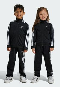 Des survêtements noirs avec des accents à trois rayures blanches, des vestes à zip et des pantalons assortis. Logos en haut à gauche de chaque veste et pantalon.