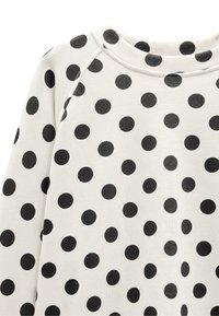 Biały sweter z dużymi czarnymi kropkami. Ma okrągły dekolt i teksturowany materiał, co podkreśla swobodny design.