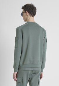 Felpa verde in cotone con collo a giro, polsi a coste e due tasche applicate sulle maniche. Tessuto liscio e vestibilità rilassata.