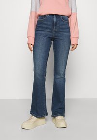 Kvinna som bär högmidjade mörkblå utsvängda jeans, en långärmad topp i rosa och grått, samt off-white plattformssneakers står på en grå golv.