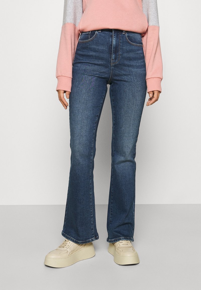 Kvinna som bär högmidjade mörkblå utsvängda jeans, en långärmad topp i rosa och grått, samt off-white plattformssneakers står på en grå golv.