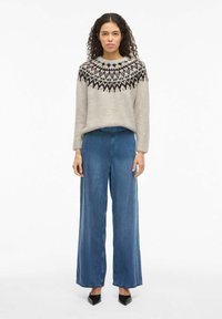 Kobieta z kręconymi włosami, ubrana w beżowy sweter w wzory, niebieskie spodnie z szerokimi nogawkami i czarne buty na obcasie, stoi na tle białym.