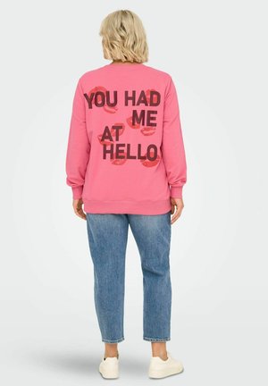 Pink sweatshirt with black text "YOU HAD ME AT HELLO" and red lip print on the back. Finished with ribbed cuffs and hem, worn with blue jeans.