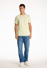 Camiseta de algodón verde claro con "COPENHAGUE" impreso en azul, combinada con unos vaqueros de mezclilla azules y zapatillas blancas. Diseño simple y casual.