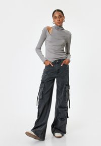 Graues langärmliges Oberteil mit asymmetrischem Ausschnitt, kombiniert mit dunklen Cargo-Hosen mit Seitentaschen und lockerer Passform. Weiße Schuhe mit strukturiertem Sohlen.
