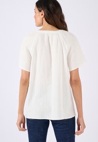Witte textuur blouse met korte mouwen en een ronde halslijn. De achterkant heeft geplooide details voor extra flexibiliteit. Gecombineerd met een donkere spijkerbroek.