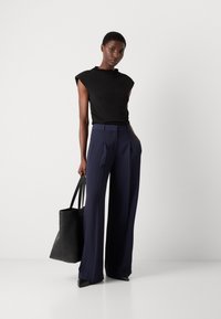 Haut noir ajusté avec des accents aux épaules, associé à un pantalon large à rayures marines. Le mannequin tient un sac tote en cuir noir et porte des escarpins pointus.
