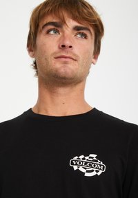 Schwarzes langärmliges T-Shirt aus Baumwolle mit Rundhalsauschnitt, das auf der Brust ein weißes Volcom-Logo im karierten Design zeigt.