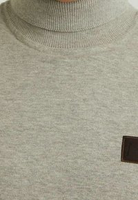 Pull à col roulé gris clair en maille douce, avec des accents côtelés au col et une petite étiquette brune sur le côté.