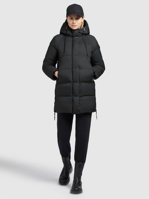 Schwarze Steppjacke mit Kapuze, einem gesteppte Design, Kordelzügen und Seitentaschen, kombiniert mit schwarzer Hose und Stiefeletten.