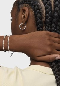 Pendientes de aro de plata y un pequeño pendiente de acento, llevados con el cabello trenzado. Una mano descansa sobre el cuello, exhibiendo una pulsera de perlas con acentos oscuros.