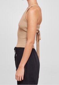 Žena v bezrukávovém béžovém žebrovaném topu s detaily na zavazování vzadu a volných černých kalhotách se stahovací šňůrkou, stojící v profilu.