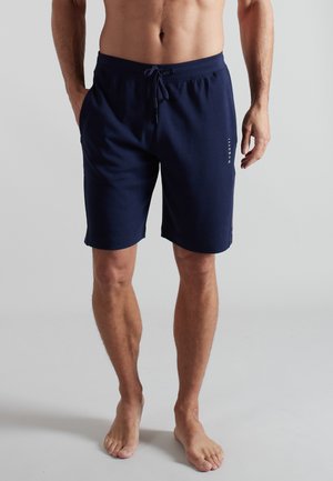 Mann trägt marineblaue Shorts mit Kordelzug, steht barfuß vor einem schlichten hellgrauen Hintergrund.