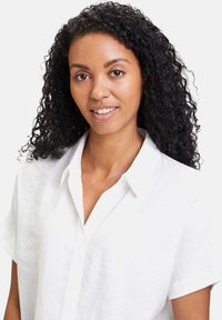 Weißes kurzärmeliges Button-Down-Hemd aus strukturiertem Stoff, mit Kragen und lockerer Passform, getragen von einer Person mit lockigem Haar.