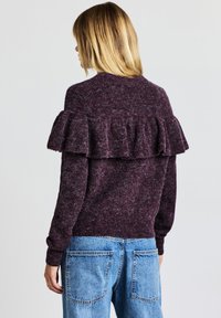 Bordeauxfarbenes Strickpullover mit gerafftem Rückendetail, langen Ärmeln und gerippten Bündchen, kombiniert mit hellblauen Jeans aus Denim mit Gesäßtaschen.
