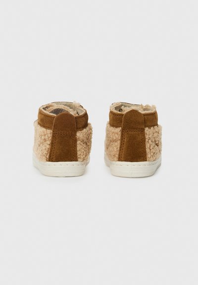Et par brune og beige fuzzy slip-on sneakers med hvite gummisåler, vist fra baksiden på en enkel hvit bakgrunn.