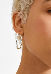 Hõbedast kõrvarõngas tekstuurse viimistlusega, millel on ebaühtlane kuju ja lihvitud pind, rippumas kõrvast.