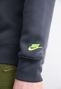 Σκούφος γκρι φούτερ με εφαρμοστή γραμμή, διακοσμημένος με φωτεινό νεον πράσινο λογότυπο Nike στον γιακά. Το ύφασμα φαίνεται λείο με ριμπ μανσέτες.