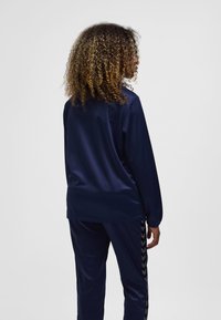 Marinblå sportsoverall, långärmad tröja med rund hals, matchande byxor med sidochevronränder, slät tygstruktur.