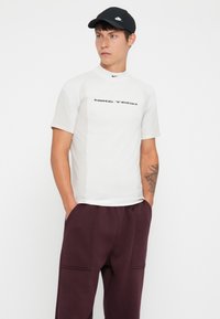 Short-sleeve white athletic shirt with "NIKE TECH" printed in black, paired with dark purple sweatpants. Materials appear soft and smooth.