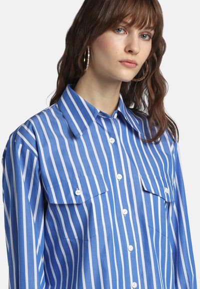 Jauna sieviete, kas valkā zilas un baltas vertikālas svītras kreklu ar pogām, kabatām ar vākiem un lielām riņķa formas auskarēm.