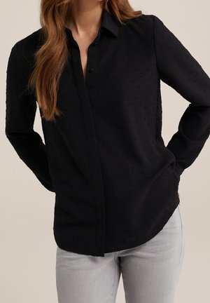 Kvinde iført en sort, tekstureret, langærmet skjorte med knapper og lysegrå bukser, stående med hænderne på hofterne mod en ensfarvet baggrund.