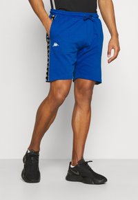 Siniset urheilushortsit, joissa on naru vyötäröllä, sivutaskut ja mustat kuviolliset sivupaneelit. Kangas on pehmeä ja rento malli.