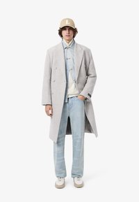 Cappotto lungo grigio chiaro in lana con design doppiopetto, indossato su una camicia di denim, jeans blu strappati e cappellino beige con logo "L". Sneakers bianche.