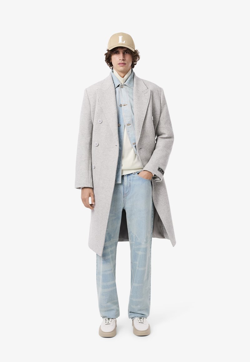 Manteau long en laine gris clair avec design à double boutonnage, porté sur une chemise en denim, un jean bleu usé et une casquette beige avec le logo "L". Baskets blanches.