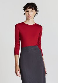 Maglietta rossa a maniche lunghe con un design aderente, abbinata a una gonna grigia scura e textured. Il tessuto appare liscio ed elegante.