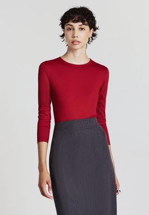 Maglietta rossa a maniche lunghe con un design aderente, abbinata a una gonna grigia scura e textured. Il tessuto appare liscio ed elegante.