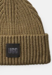 Căciulă tricotată în verde măsliniu cu textură cu nervuri. Prezintă un patch rectangular negru embosat cu "UGG" în față.