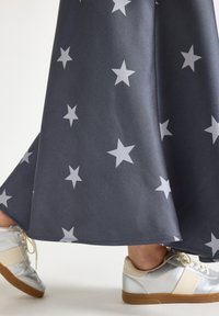 Jambes inférieures dans des baskets argentées, avançant, portant une jupe sombre avec des motifs d'étoiles clairs, sur un fond blanc uni.