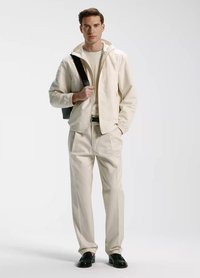 Mężczyzna w kremowej kurtce i spodniach z czarnym paskiem, czarnymi butami oraz czarną torbą na ramię, stojący na białym tle.