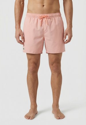 Hombre que lleva bañador corto de color rosa claro con cintura elástica y cordón, de pie descalzo frente a un fondo liso.