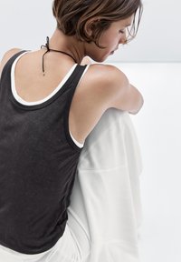 Femme aux cheveux courts portant un débardeur noir et un pantalon blanc, assise avec les genoux relevés, regardant vers le bas dans un cadre clair et minimaliste.