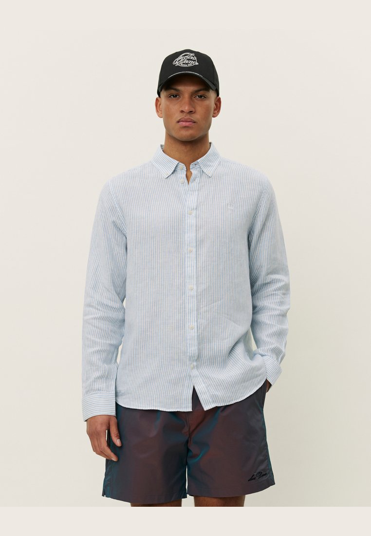 Camicia a righe blu chiaro con bottoni, realizzata in un tessuto dalla superficie liscia, abbinata a pantaloni corti scuri. L'individuo indossa un cappellino nero.