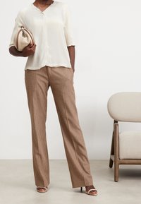 Blouse crème à manches courtes avec un décolleté en V, associée à un pantalon large à carreaux marron. Le mannequin tient un sac à main beige et porte des sandales beiges.