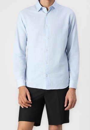 Mężczyzna ubrany w jasnoniebieską koszulę z długim rękawem zapinaną na guziki oraz czarne spodenki do kolan na jednolitym tle.