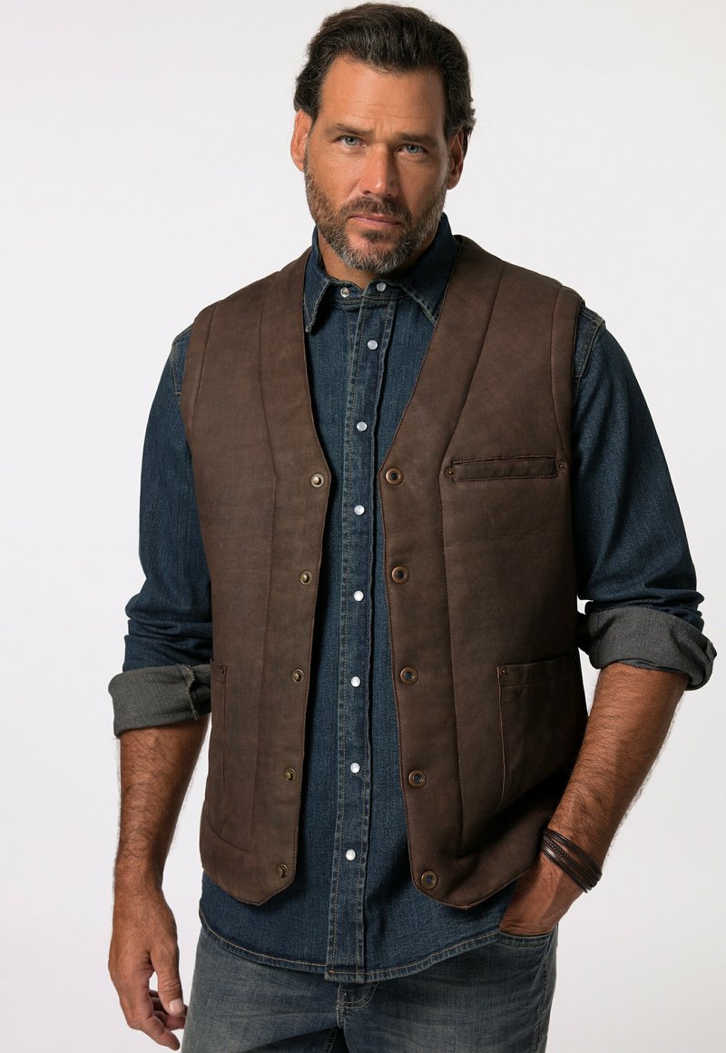 Gilet en cuir marron avec une fermeture à boutons, comportant une poche poitrine et deux poches avant. Porté sur une chemise en denim aux manches retroussées.