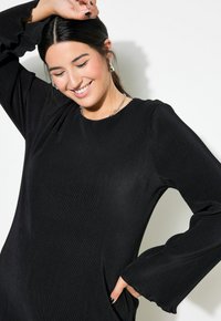 Zwarte geribde jurk met lange, uitlopende mouwen. Heeft een ronde halslijn en een zachte textuur. Model glimlachend, toont sieraden en haardos.