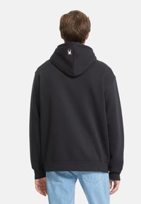 Felpa nera con una texture morbida, dotata di un logo sulla parte posteriore del cappuccio. Maniche a costine e orlo, vestibilità rilassata, indossata con jeans azzurri chiari.