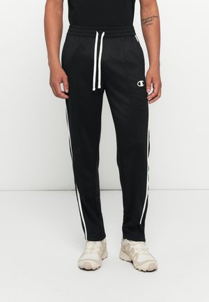 Personne portant un pantalon de sport noir avec des rayures blanches sur les côtés, des cordons blancs, et des baskets couleur crème, se tenant devant un fond blanc uni.