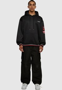 Schwarzer Hoodie mit Fronttasche, weißem Branding und roten Akzenten. Kombiniert mit baggy schwarzen Cargo-Hosen und weißen Sneakern.