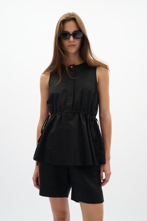 Mujer con camiseta negra sin mangas con cordón en la cintura, pantalones cortos negros y gafas de sol oscuras, posando sobre un fondo blanco liso.