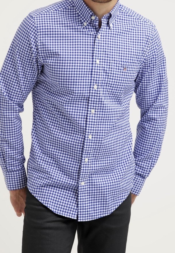 Homme portant une chemise à carreaux bleue et blanche à manches longues et un pantalon noir, debout devant un fond blanc uni.